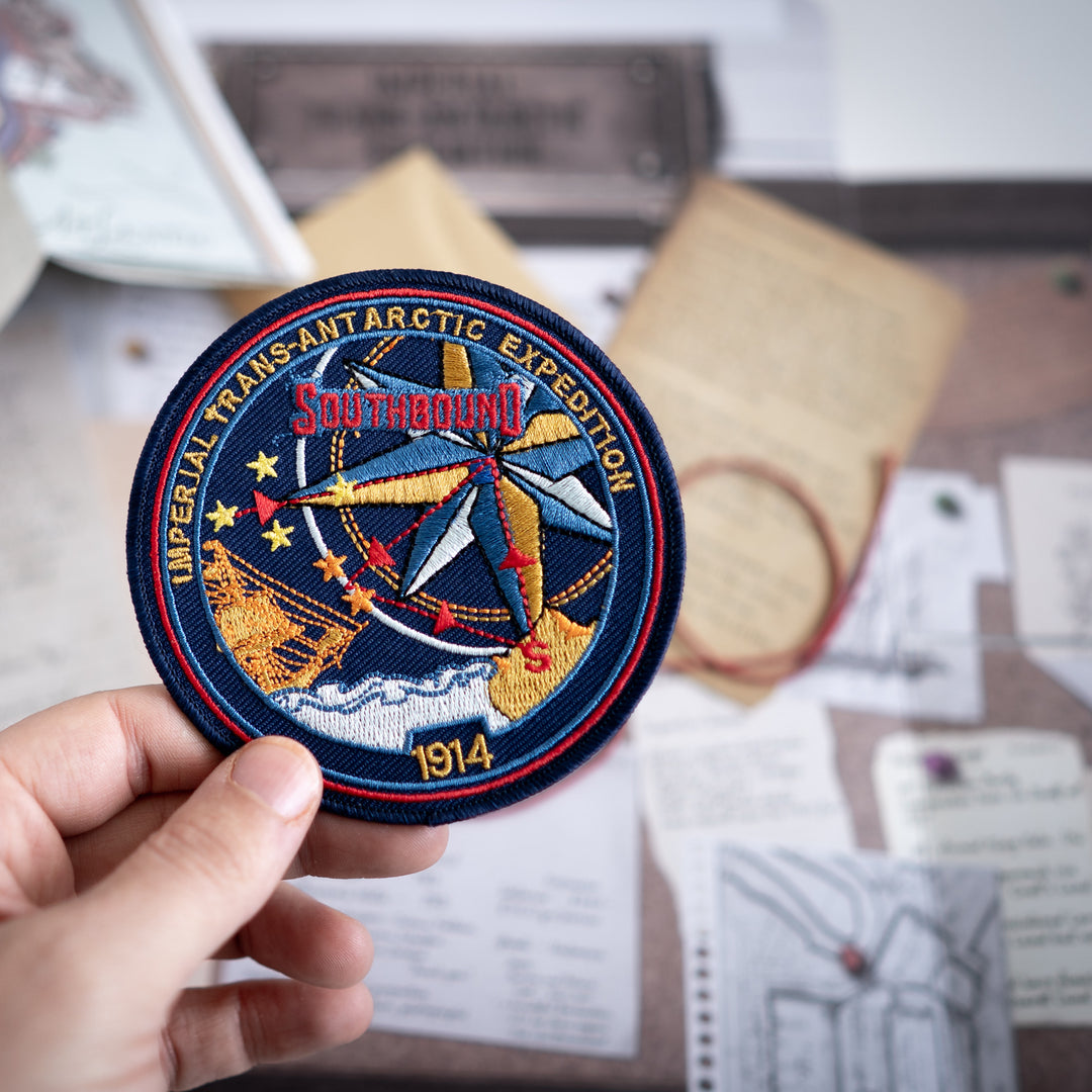 Embroidered patch of the Imperial Trans-Antarctic Expedition held by a hand on a blurred background. Games Night in an envelope and tabletop Escape Room Game. Similar to an Exit Game. Same genre as murder mystery.