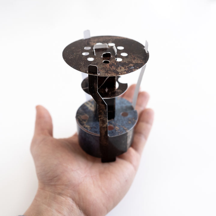 Hand holding a metallic primus stove replica. Games Night in an envelope and tabletop Escape Room Game. Similar to an Exit Game. Same genre as murder mystery.