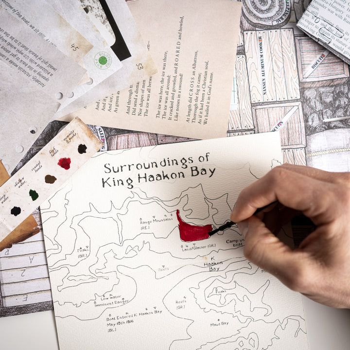 Hand coloring a map of King Haakon Bay with a red paint on a table with other maps and materials. Games Night in an envelope and tabletop Escape Room Game. Similar to an Exit Game. Same genre as murder mystery.